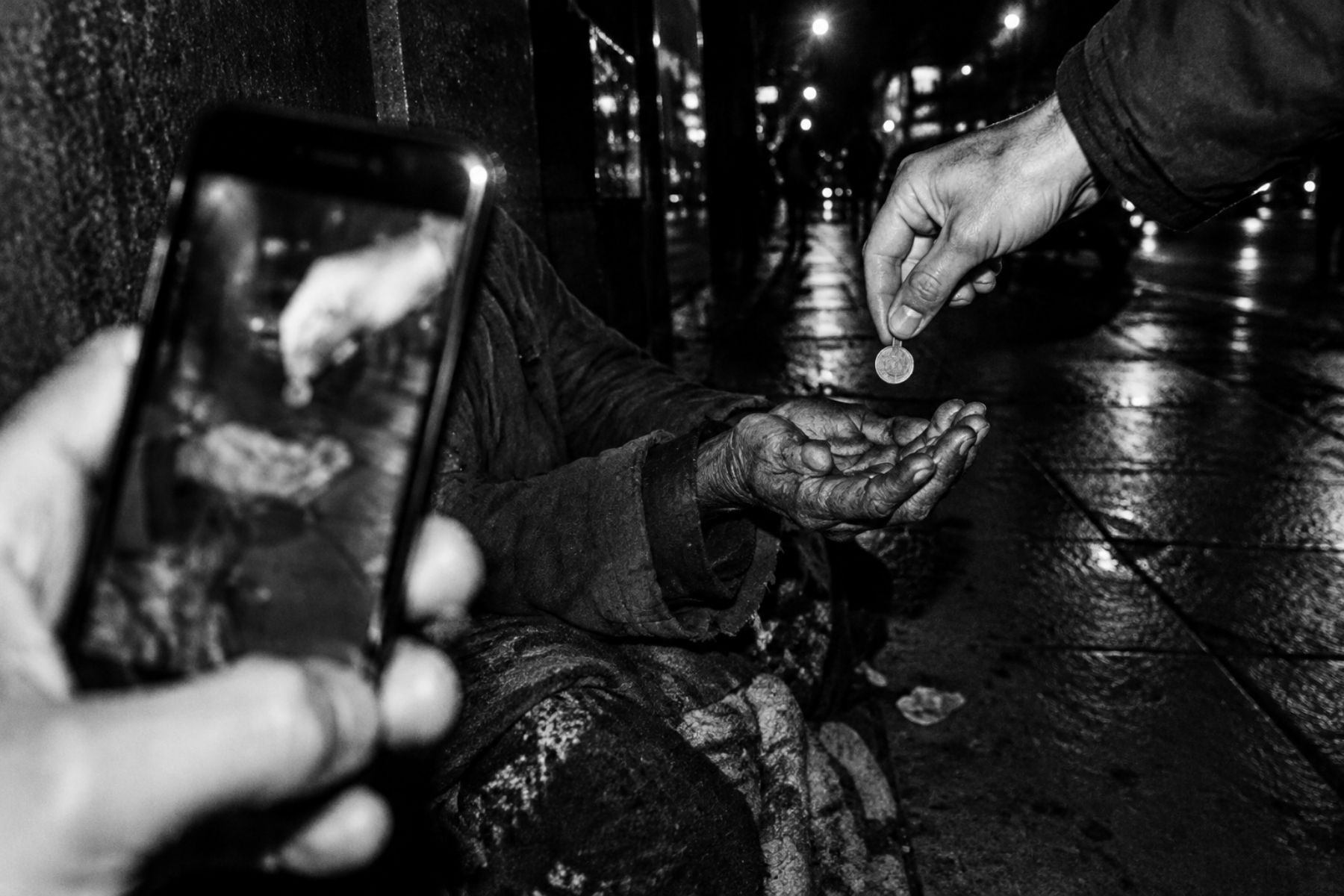 Black and white photo: a phone films someone dropping a coin into a homeless person's hand
