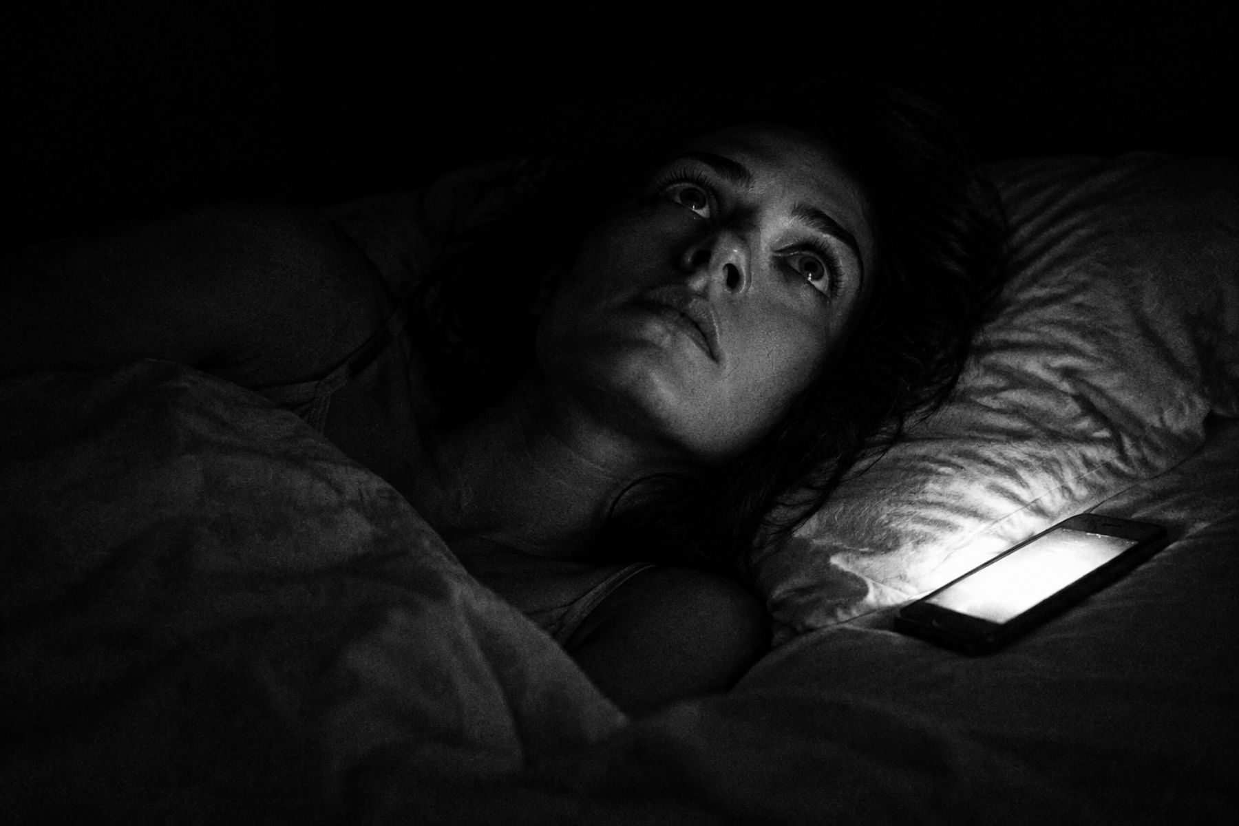 Black and white photo: a woman lying awake at night lit by her phone