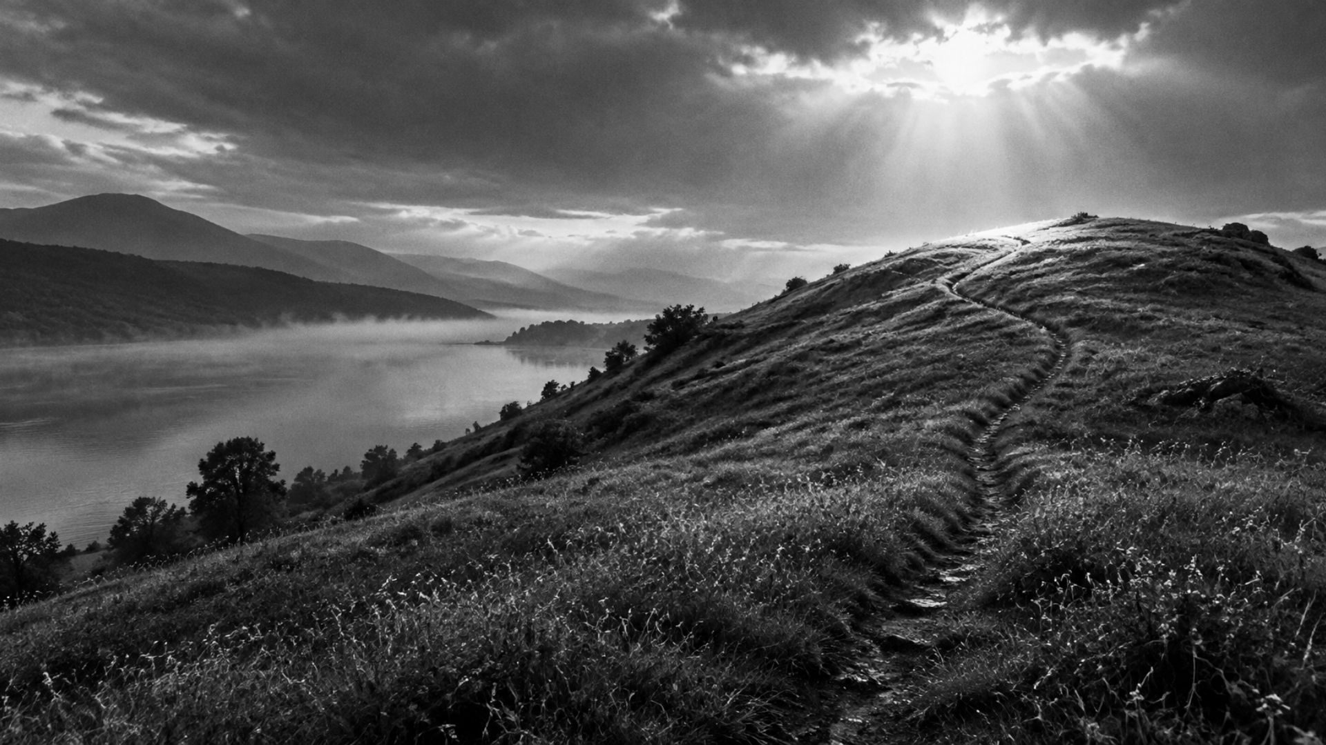 Black and white photo: an empty hillside above a lake at dawn, a worn path leading up